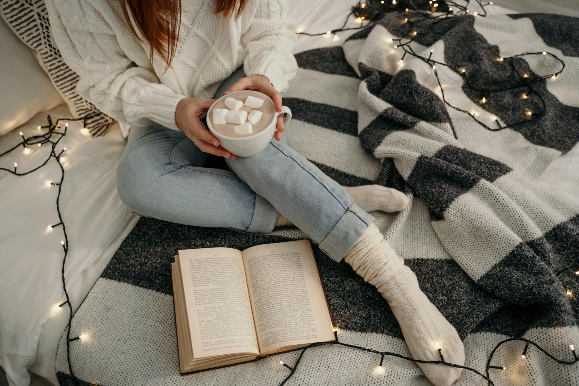 A woman is sitting on a bed holding a cup of hot chocolate and a book.