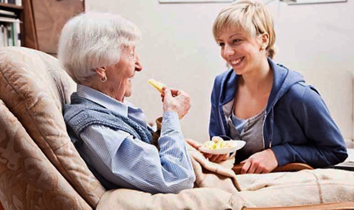 Eine Frau füttert eine ältere Person in einem Haus mit einem Stück Essen; beide lächeln.