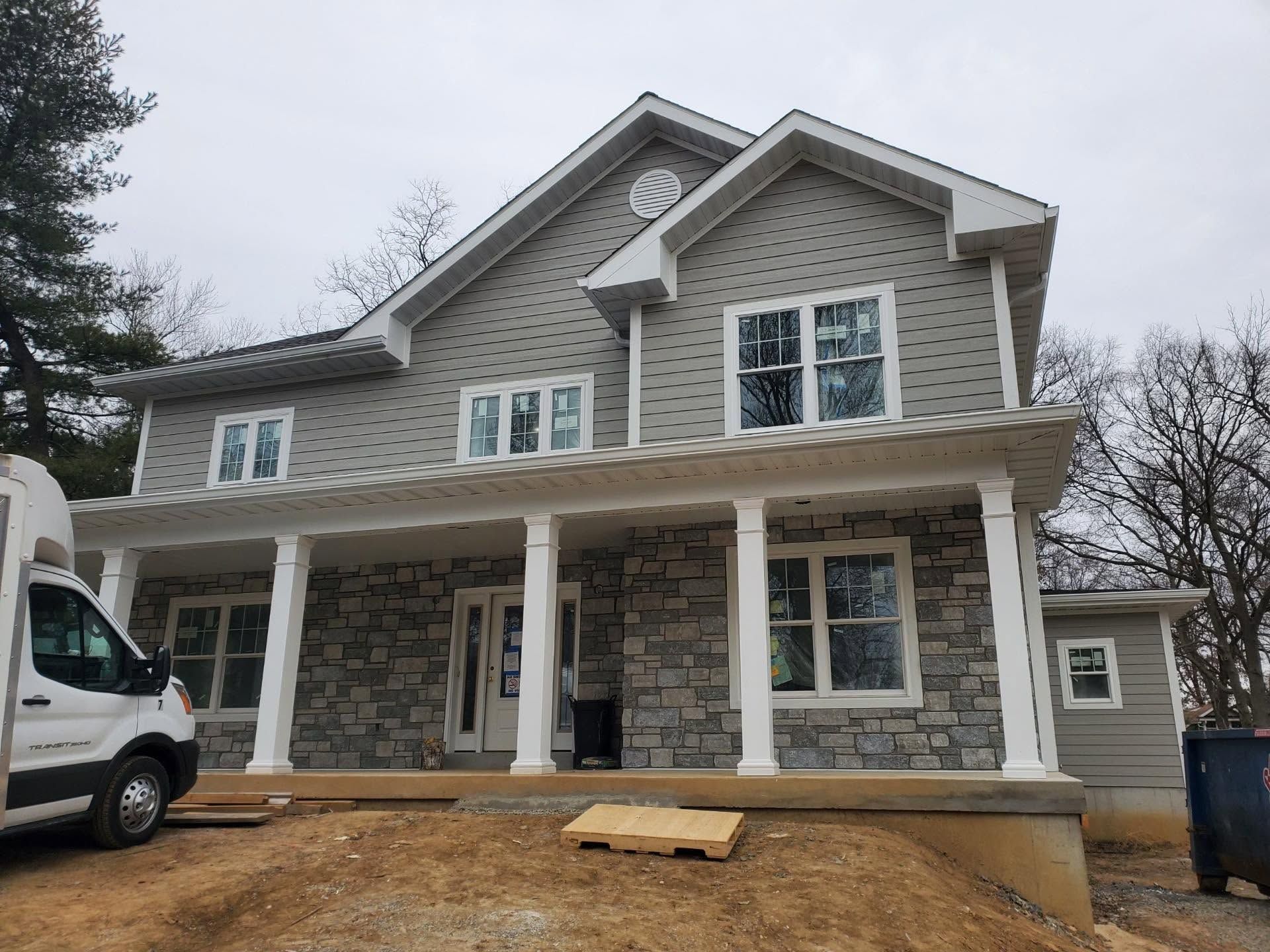 Two-story house under construction with stone and beige siding, front porch, and construction materials in front Two-story house under construction with stone and beige siding, front porch, and construction materials in front