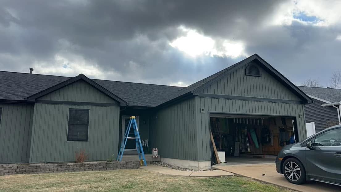 Single-story green house with an open garage, blue ladder, and a car parked in the driveway under cloudy skies Single-story green house with an open garage, blue ladder, and a car parked in the driveway under cloudy skies