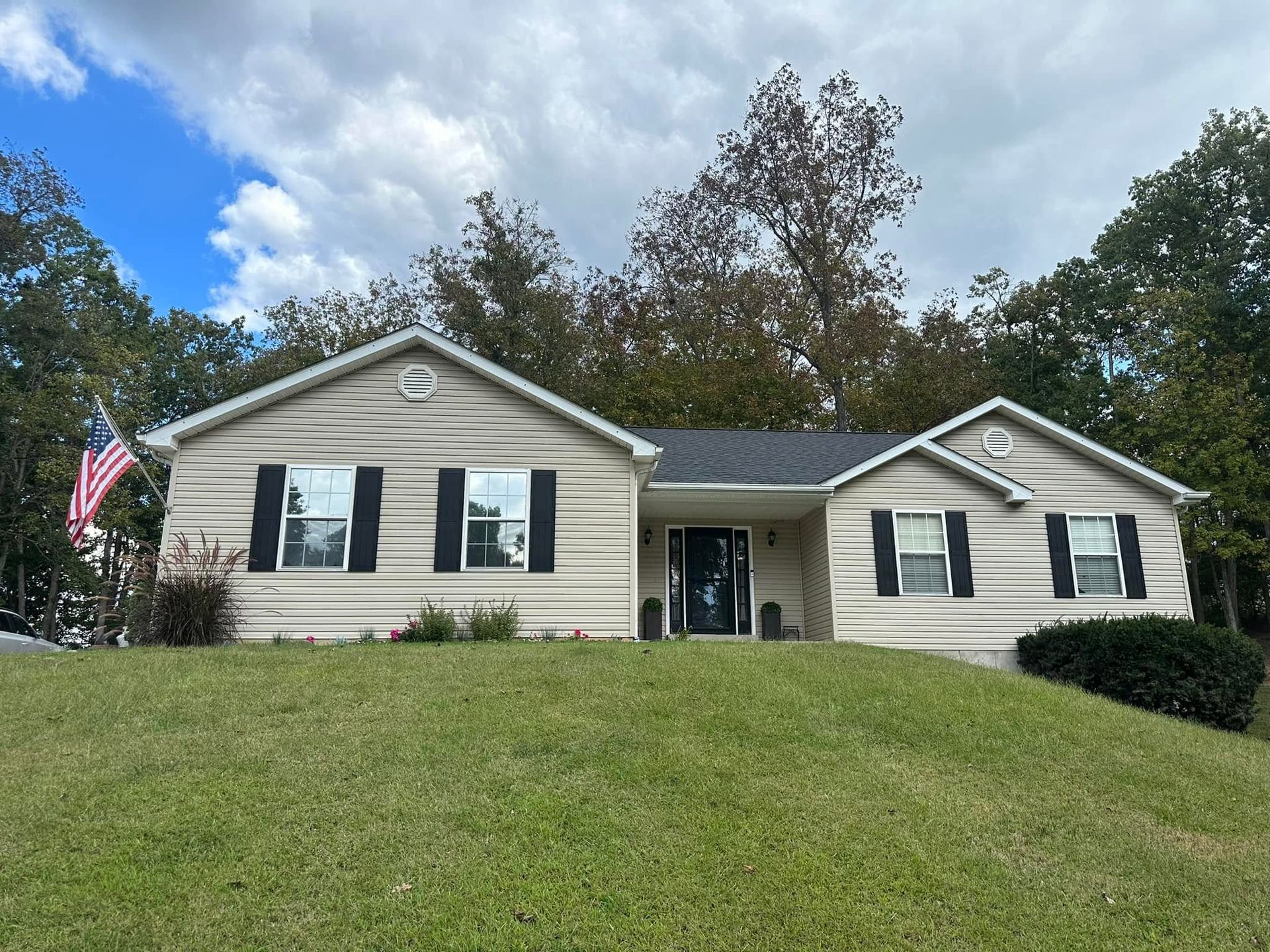 Single-story beige house with black shutters, front lawn, and an American flag under a partly cloudy sky Single-story beige house with black shutters, front lawn, and an American flag under a partly cloudy sky
