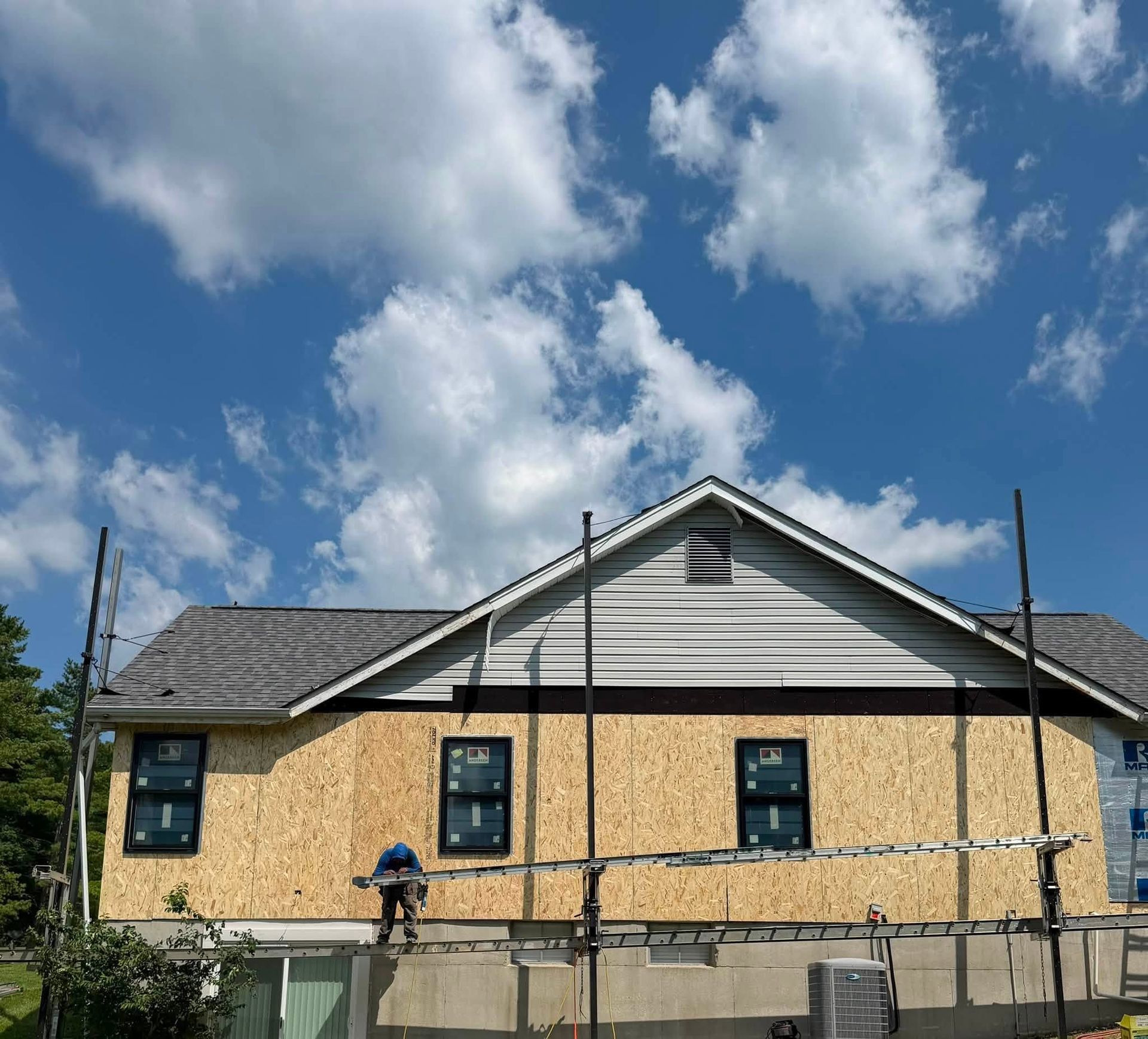 House exterior under construction with plywood siding, white gable trim, and a worker on scaffolding under a blue sky House exterior under construction with plywood siding, white gable trim, and a worker on scaffolding under a blue sky