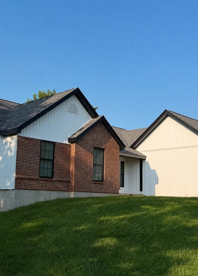 Suburban house with brick and white siding, dark roof, and green lawn under a clear blue sky Suburban house with brick and white siding, dark roof, and green lawn under a clear blue sky
