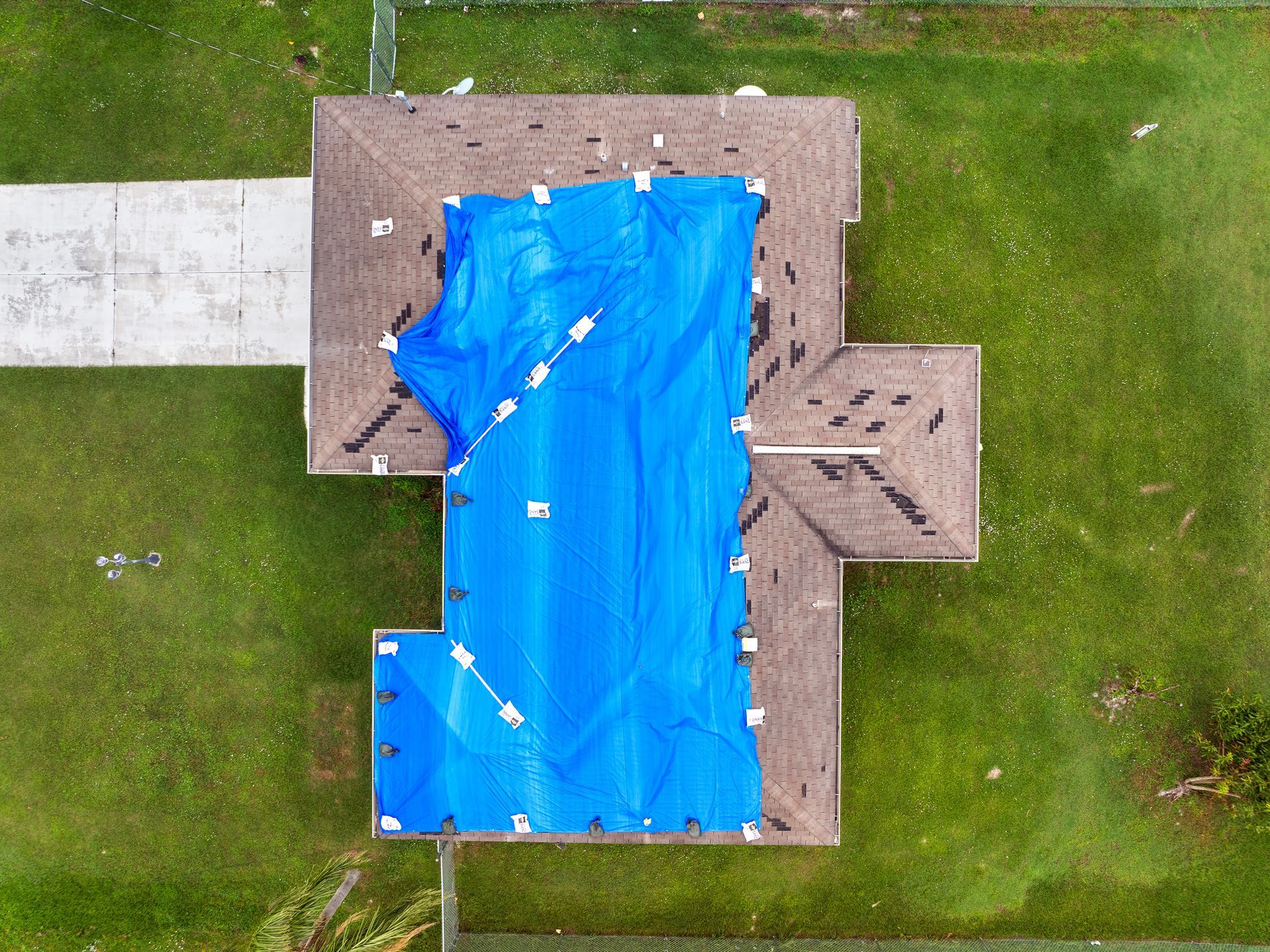 Aerial view of a house roof covered with a large blue tarp and surrounding green lawn. Aerial view of a house roof covered with a large blue tarp and surrounding green lawn.