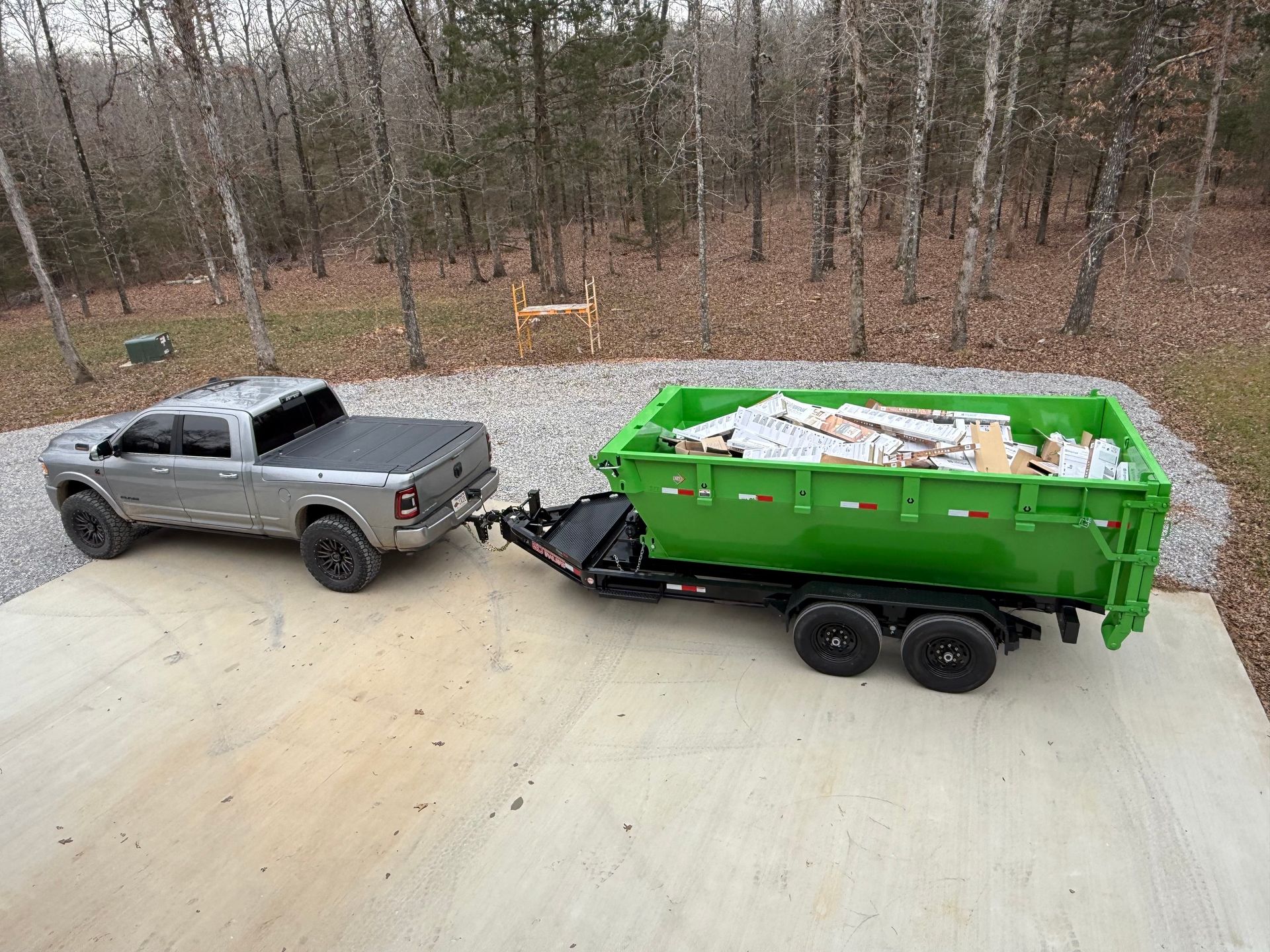 Silver pickup truck towing a green dump trailer loaded with debris on a concrete driveway by woods