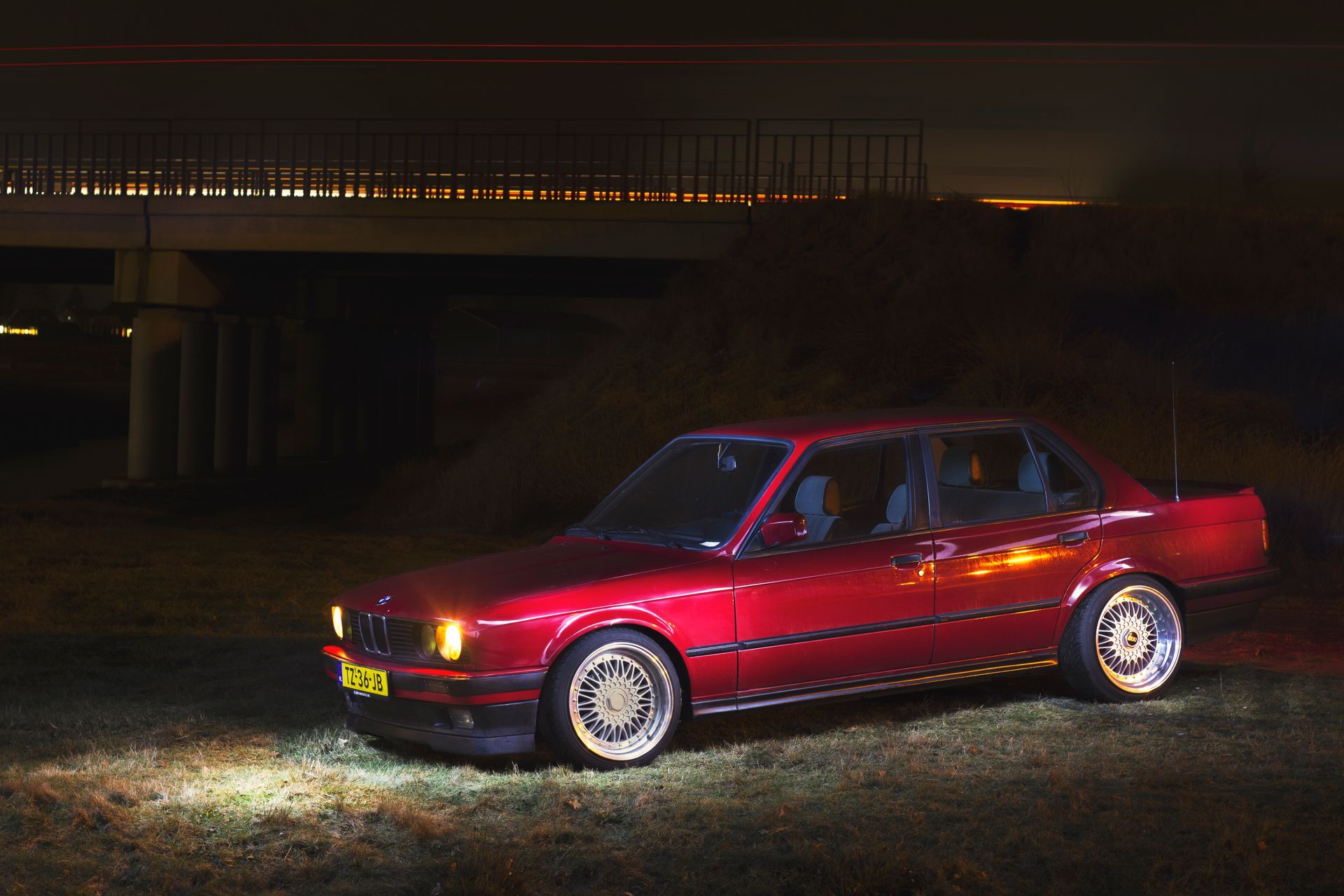 Een rode vintage BMW sedan staat 's nachts geparkeerd onder een brug, verlicht door koplampen.