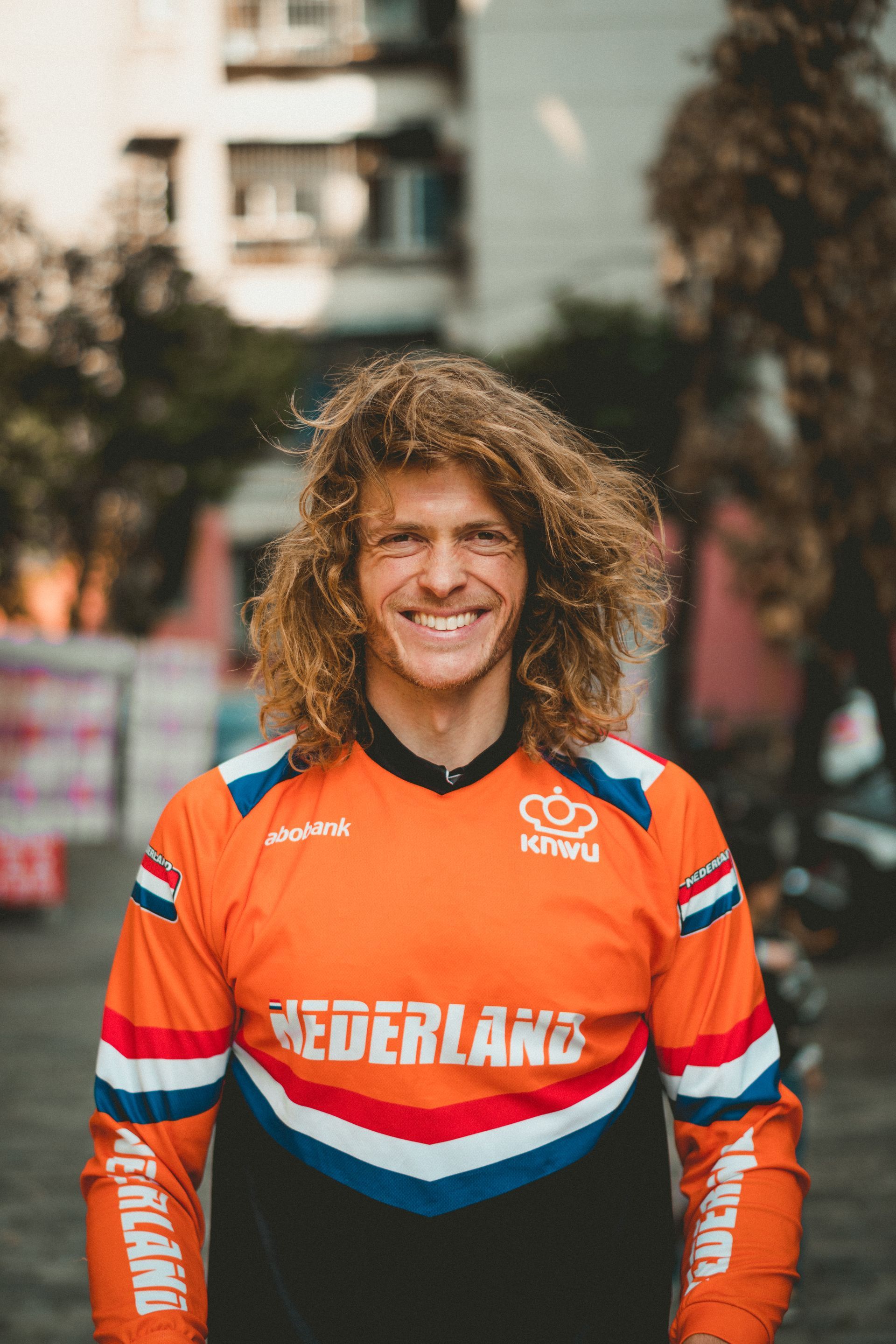 Een man met krullend haar lacht, draagt ​​een oranje 