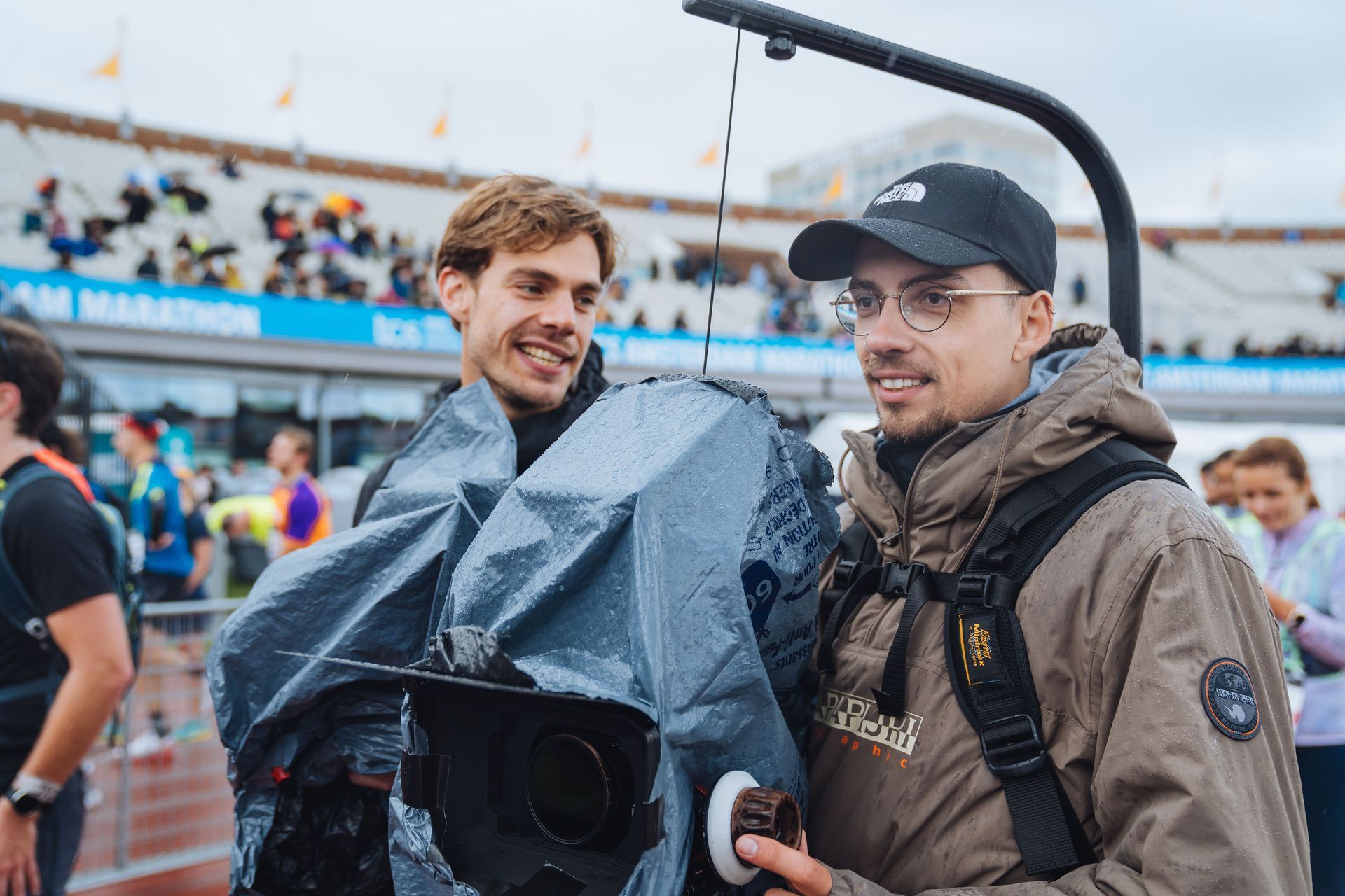 Twee mannen glimlachen naast een camera die met een zeil is afgedekt, tijdens een marathon. Een van hen draagt ​​een zwarte pet.