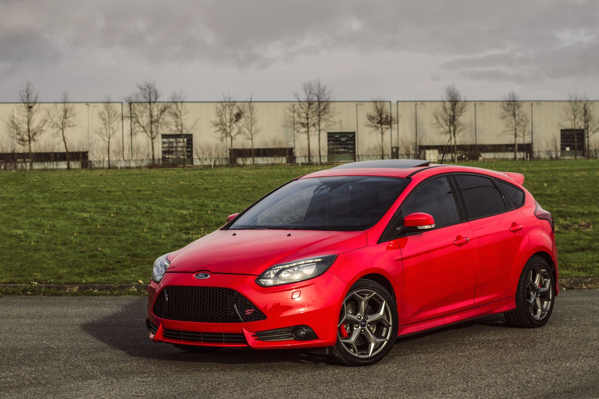 Een rode Ford Focus ST staat geparkeerd op asfalt, met groen gras en een lange, beige muur op de achtergrond.