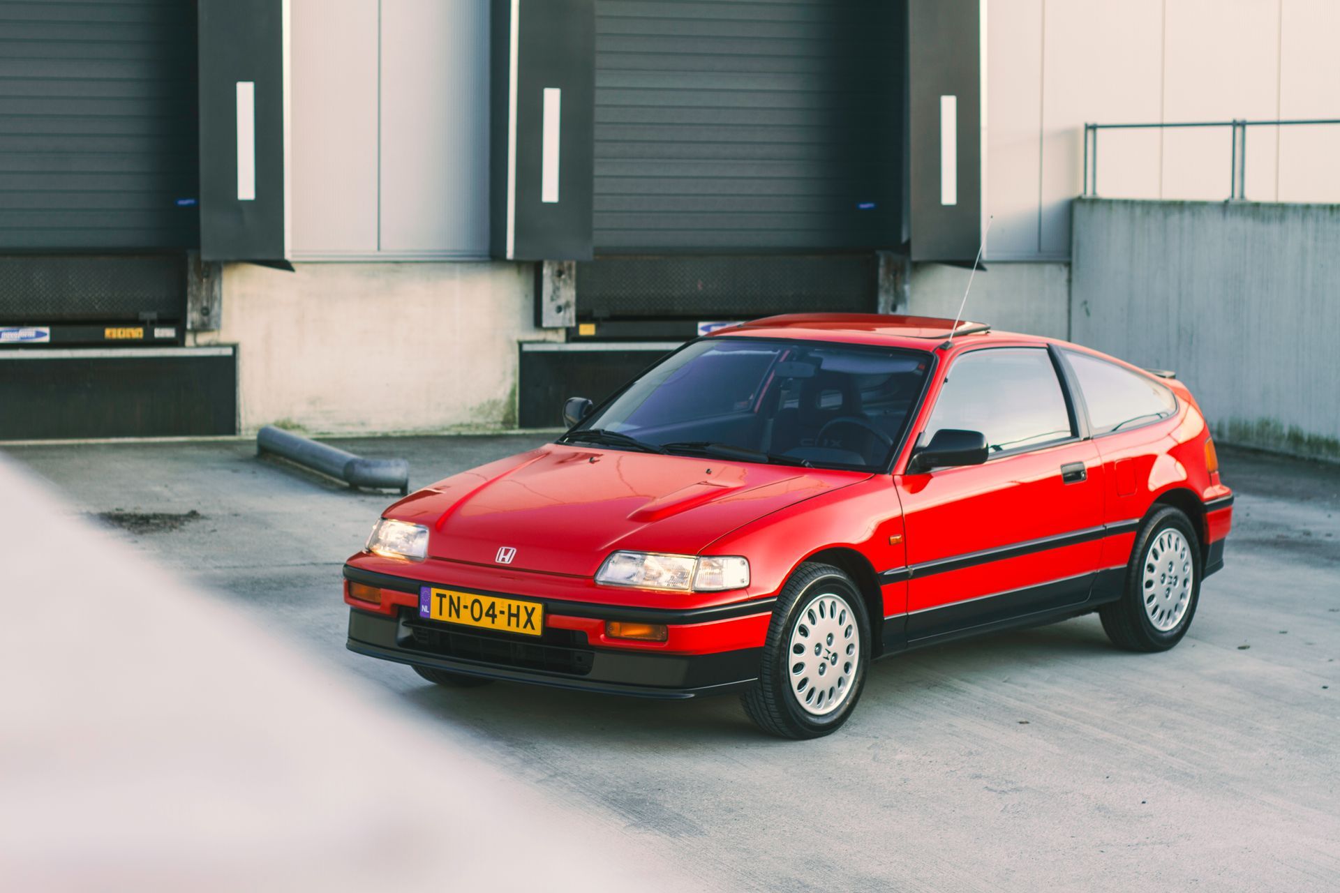 Een rode Honda CRX staat geparkeerd voor een gebouw met laadperrons.