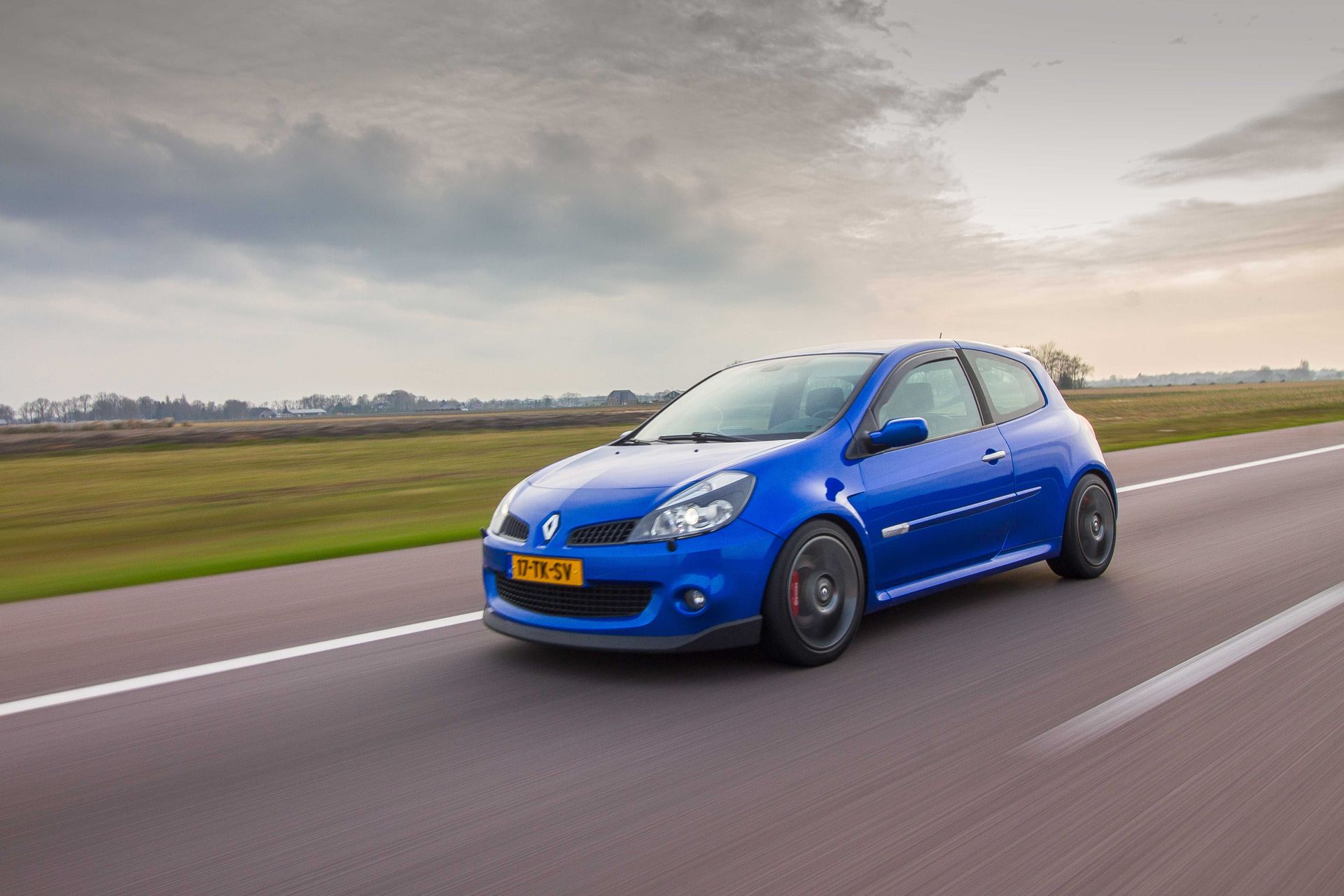 Een blauwe Renault Clio sportwagen rijdt op een weg, naar links, onder een bewolkte hemel.