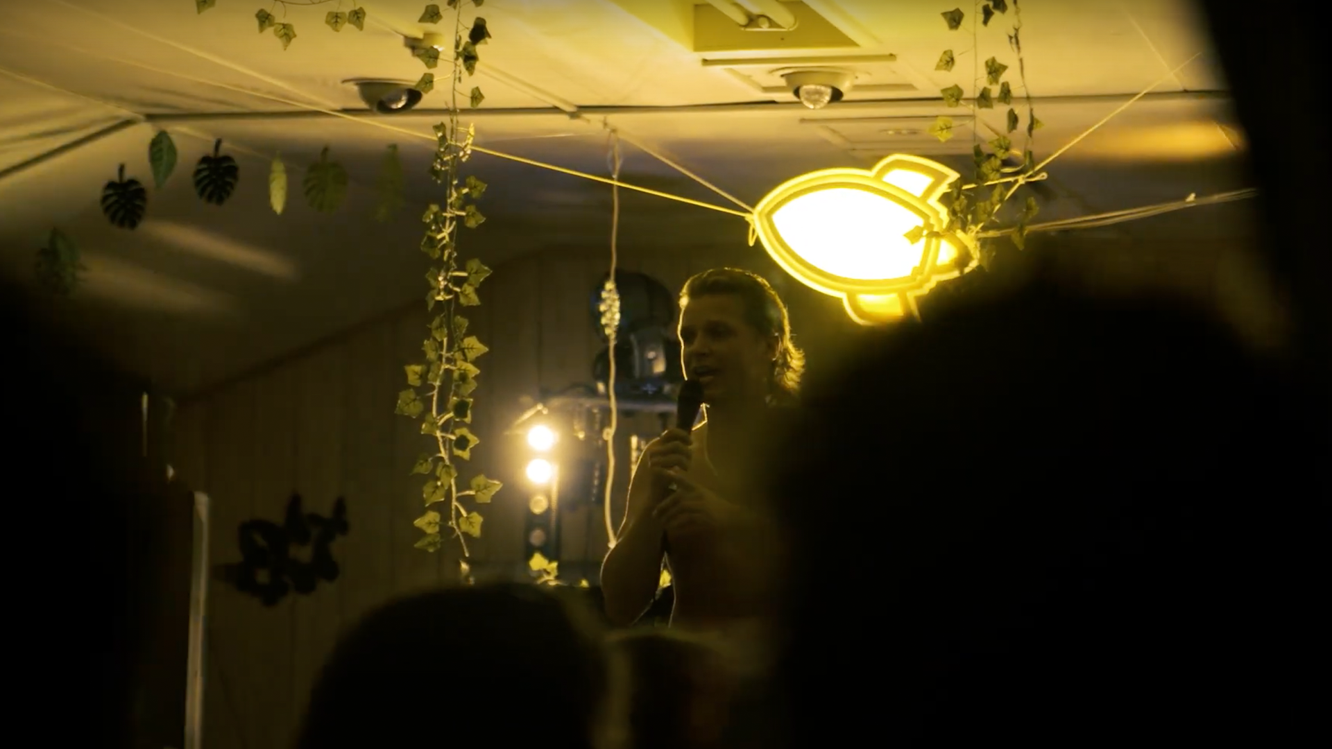 Een man spreekt in een microfoon op een podium, verlicht door spotlights en een raketvormige lichtarmatuur.