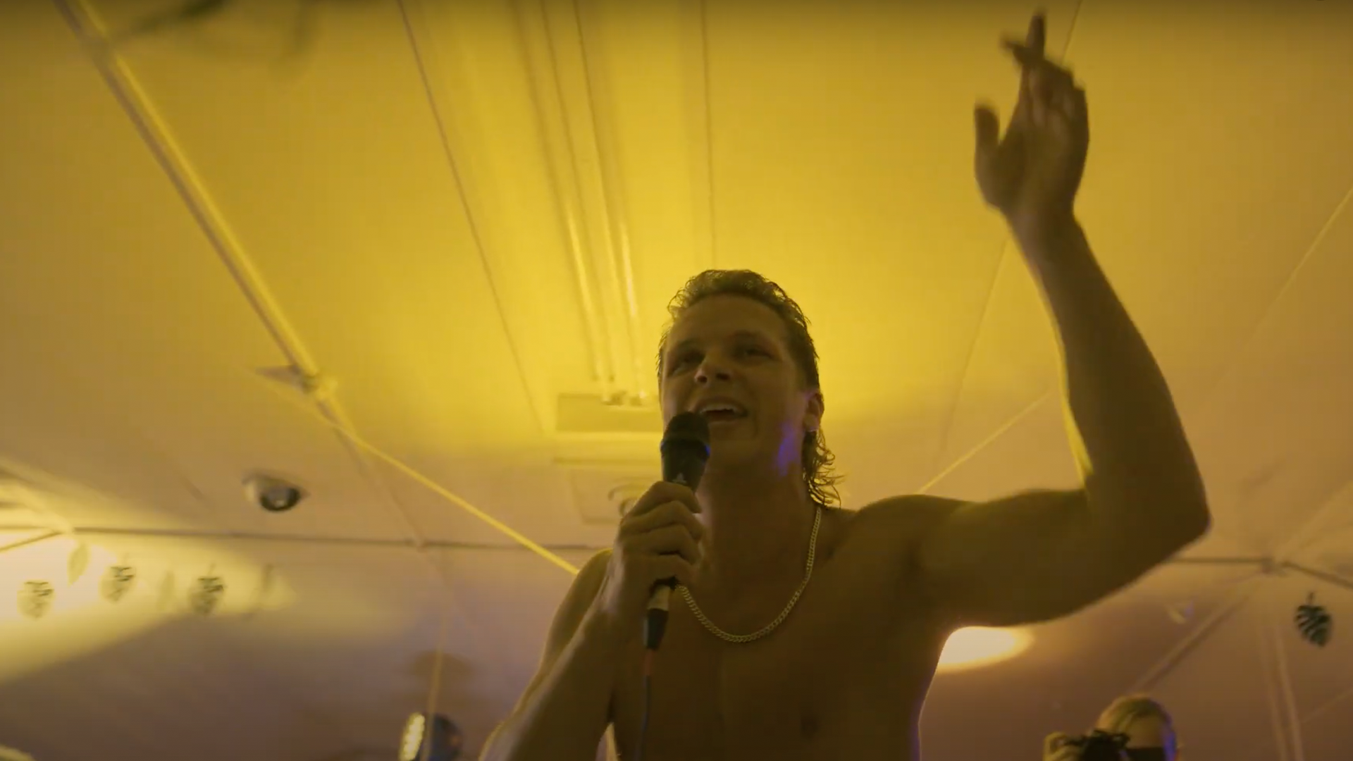 Een man met ontbloot bovenlijf, een microfoon in zijn hand en zijn arm omhoog, spreekt onder geel licht.