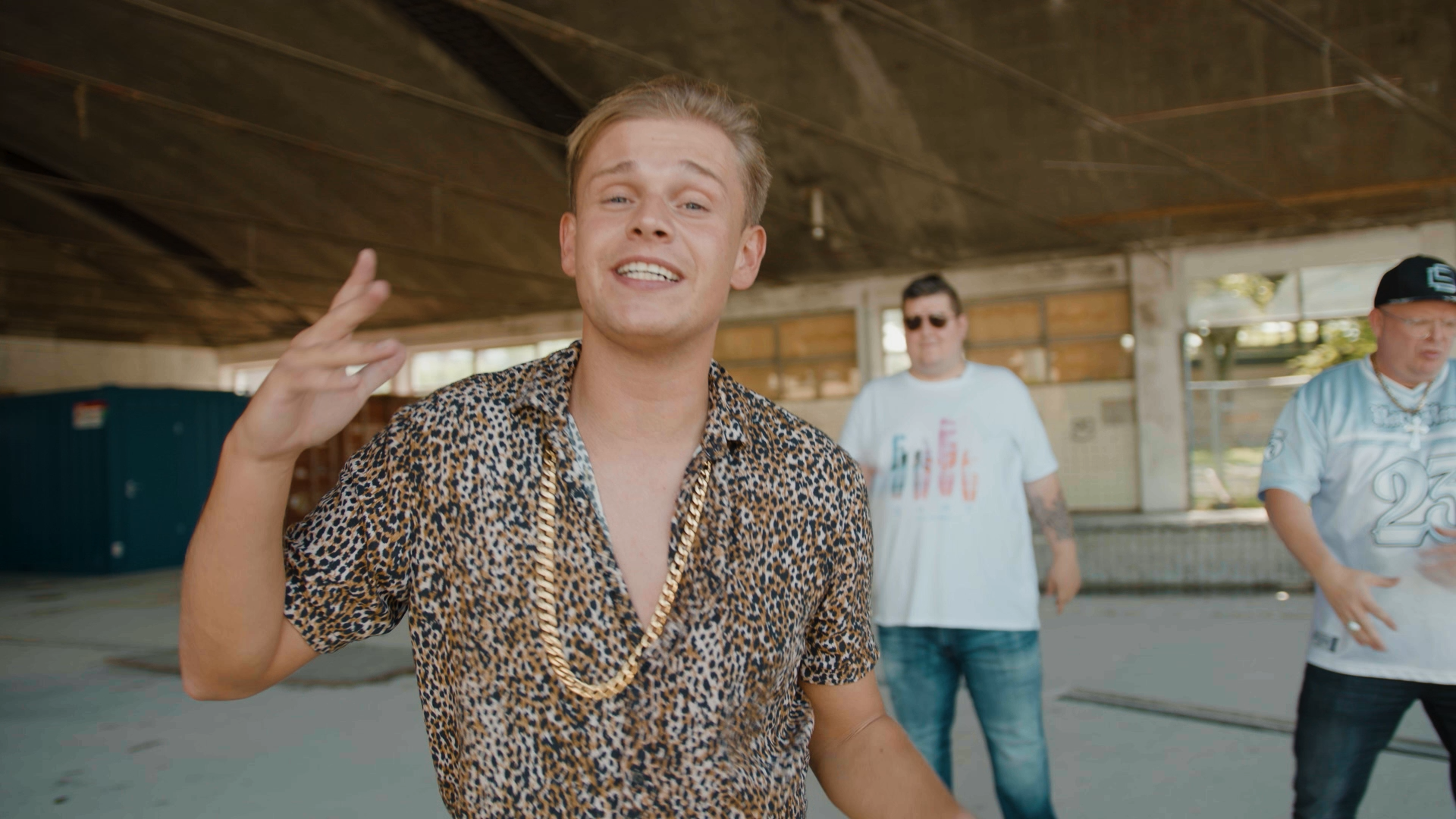 Een man in een shirt met luipaardprint en een gouden ketting maakt een vredesteken, terwijl twee mannen dansen in een vervallen gebouw.