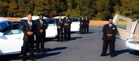 Men in suits standing near white limousines, possibly for a funeral service.