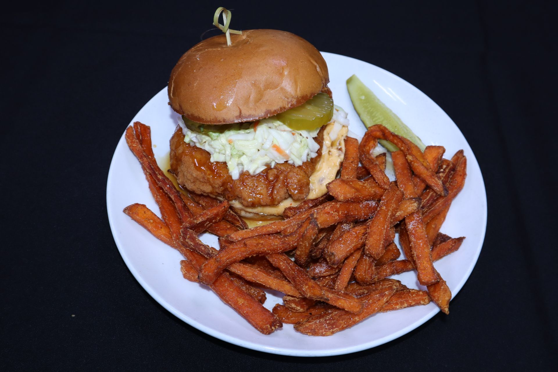 Fried chicken sandwich with coleslaw, pickles, and sweet potato fries on a plate.