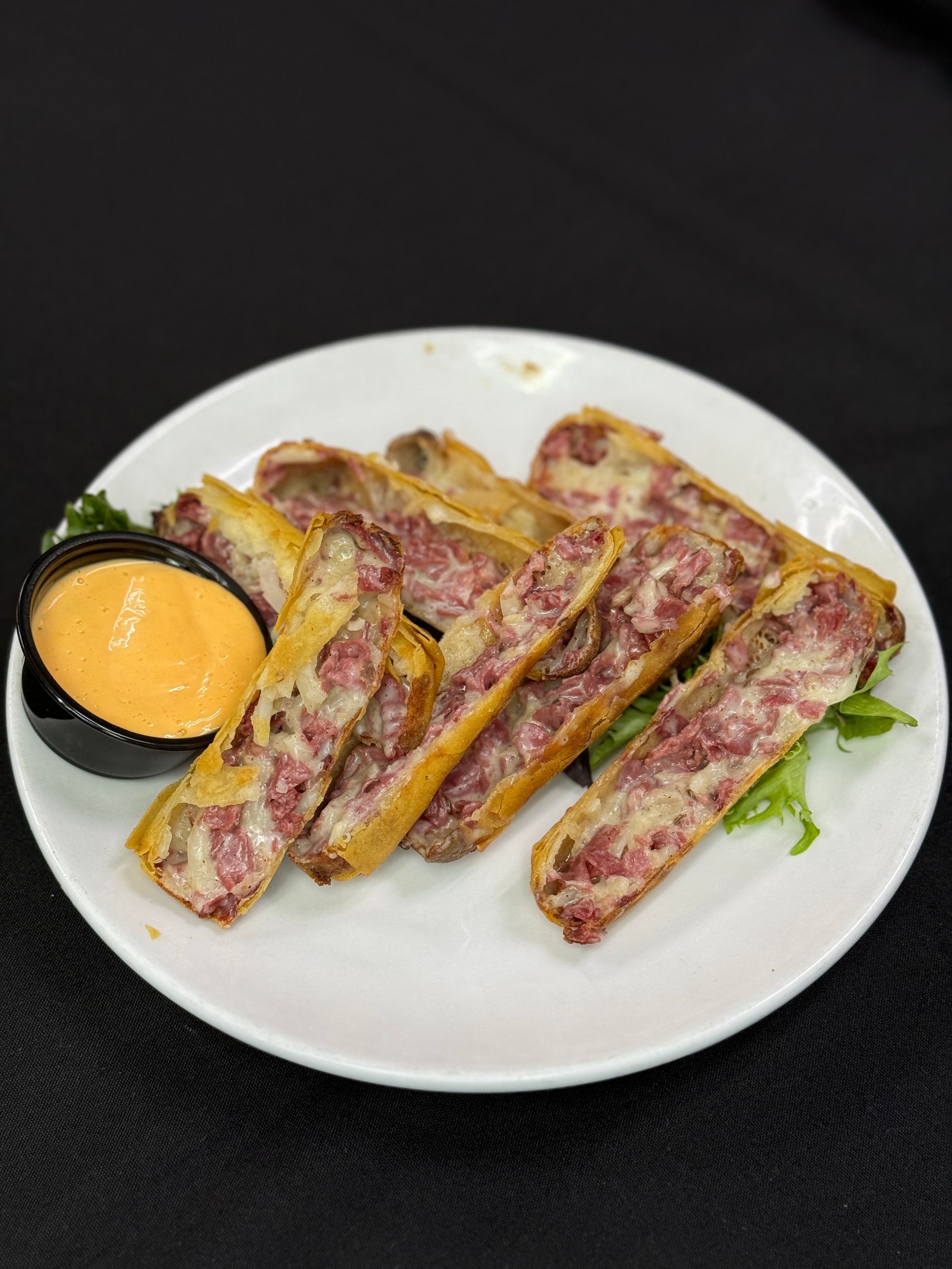 Fried egg rolls on white plate with orange dipping sauce, black background.