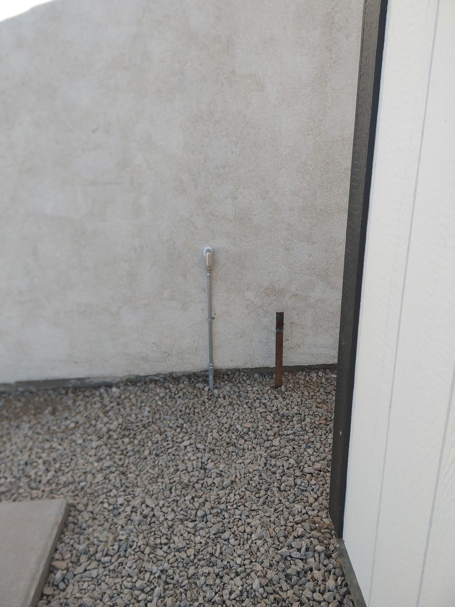 A gravel-covered yard with a wall and a shed. Two pipes extend from the ground.