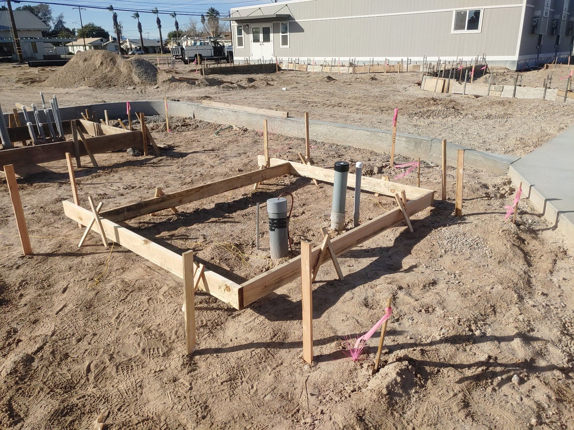 Construction site: wooden forms outline a small square foundation, with plumbing pipes and stakes in dirt.