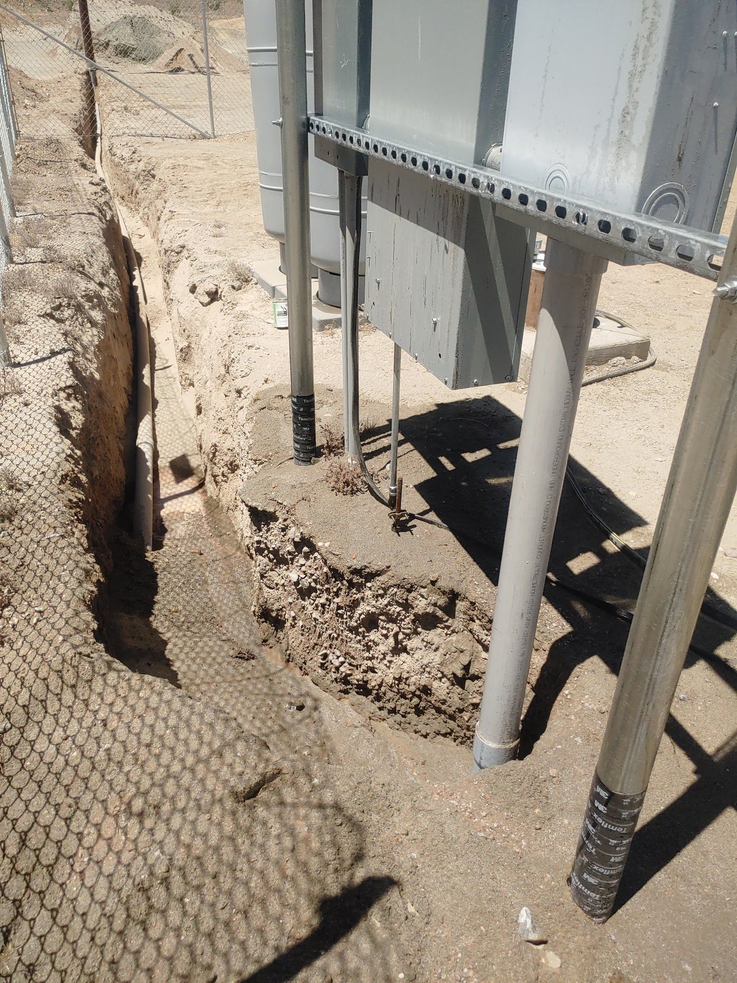 Trench next to a chain-link fence and electrical equipment. Gray poles support metal boxes on a sandy ground.
