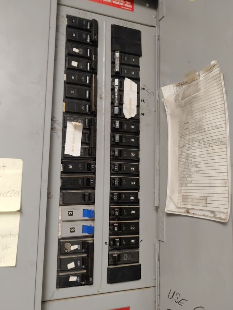 Electrical circuit breaker panel with multiple switches, inside a gray metal cabinet, with paperwork attached.
