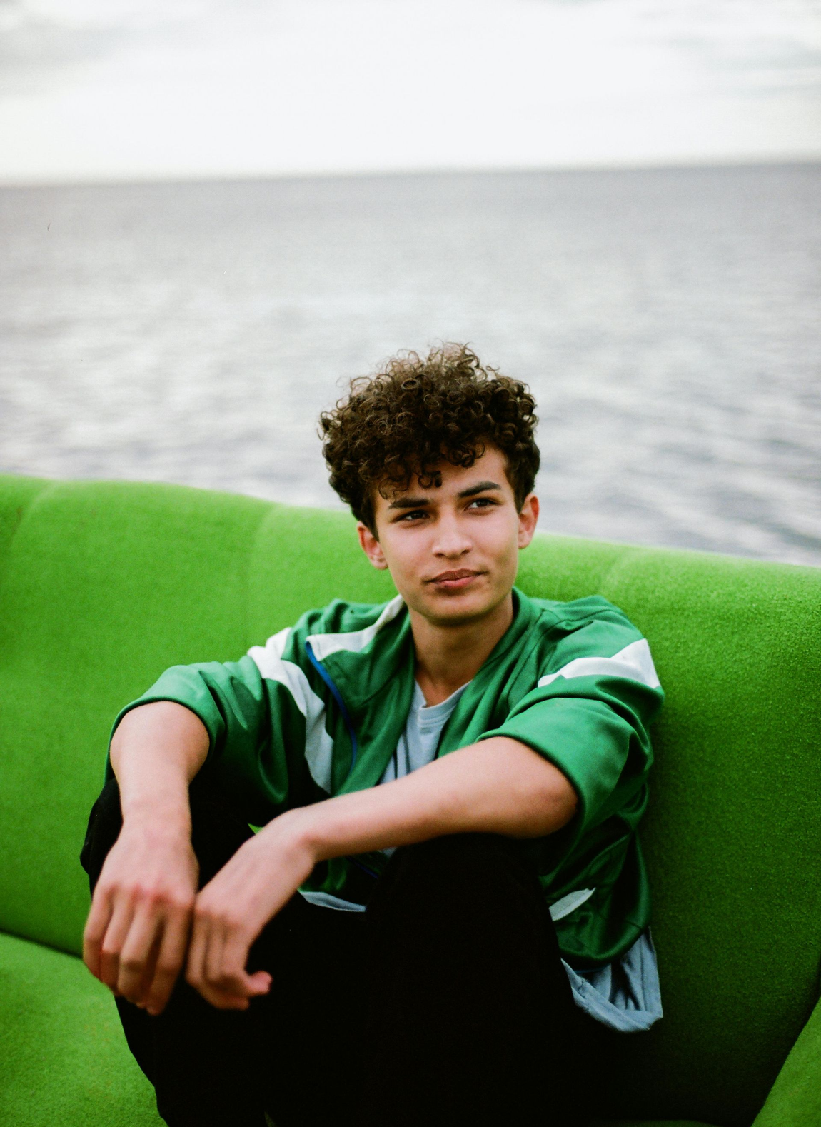 Young person sitting on a green couch, facing forward, by the sea. They wear a green jacket and black pants.