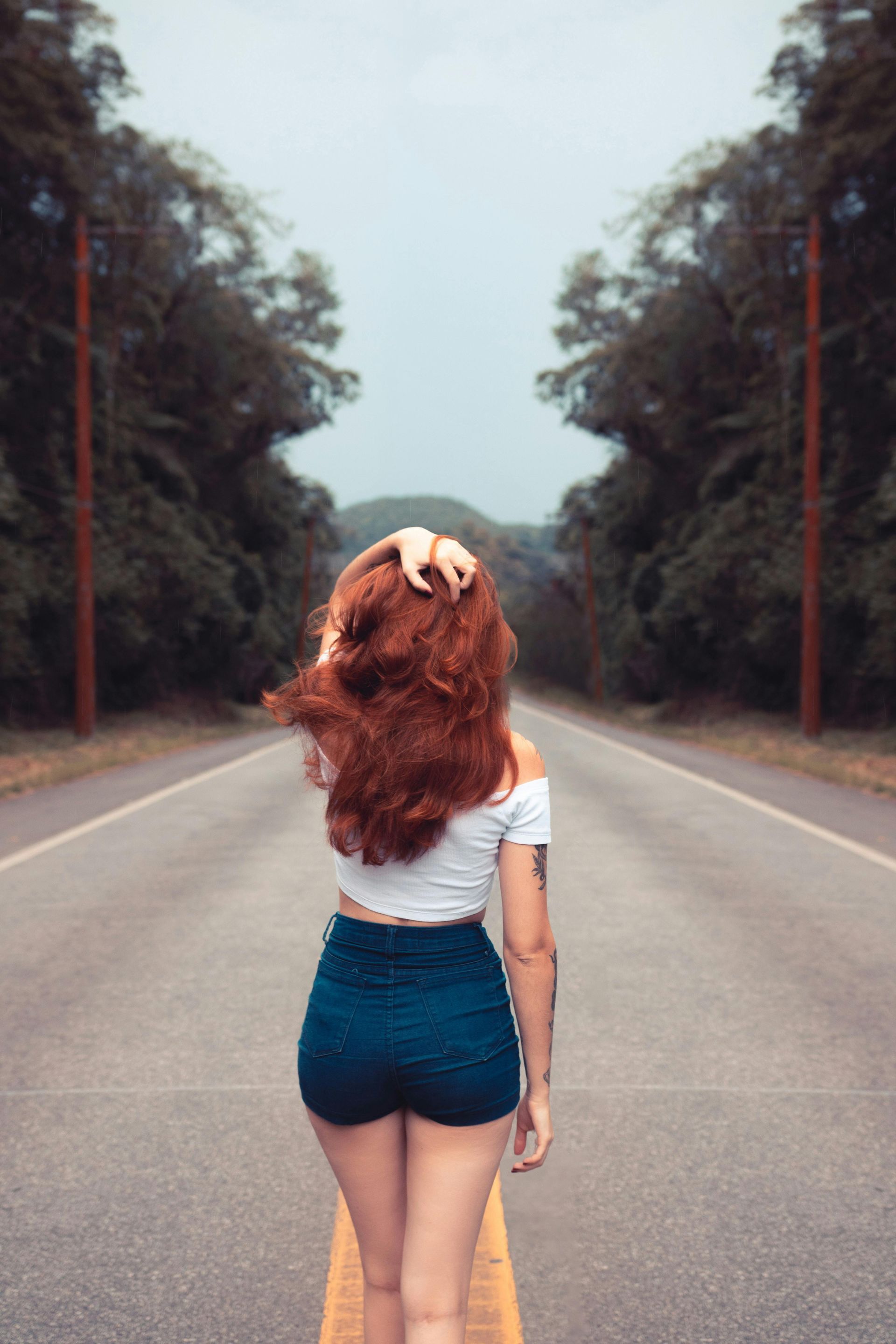 Woman with red hair and blue shorts walks down an empty road lined with trees.
