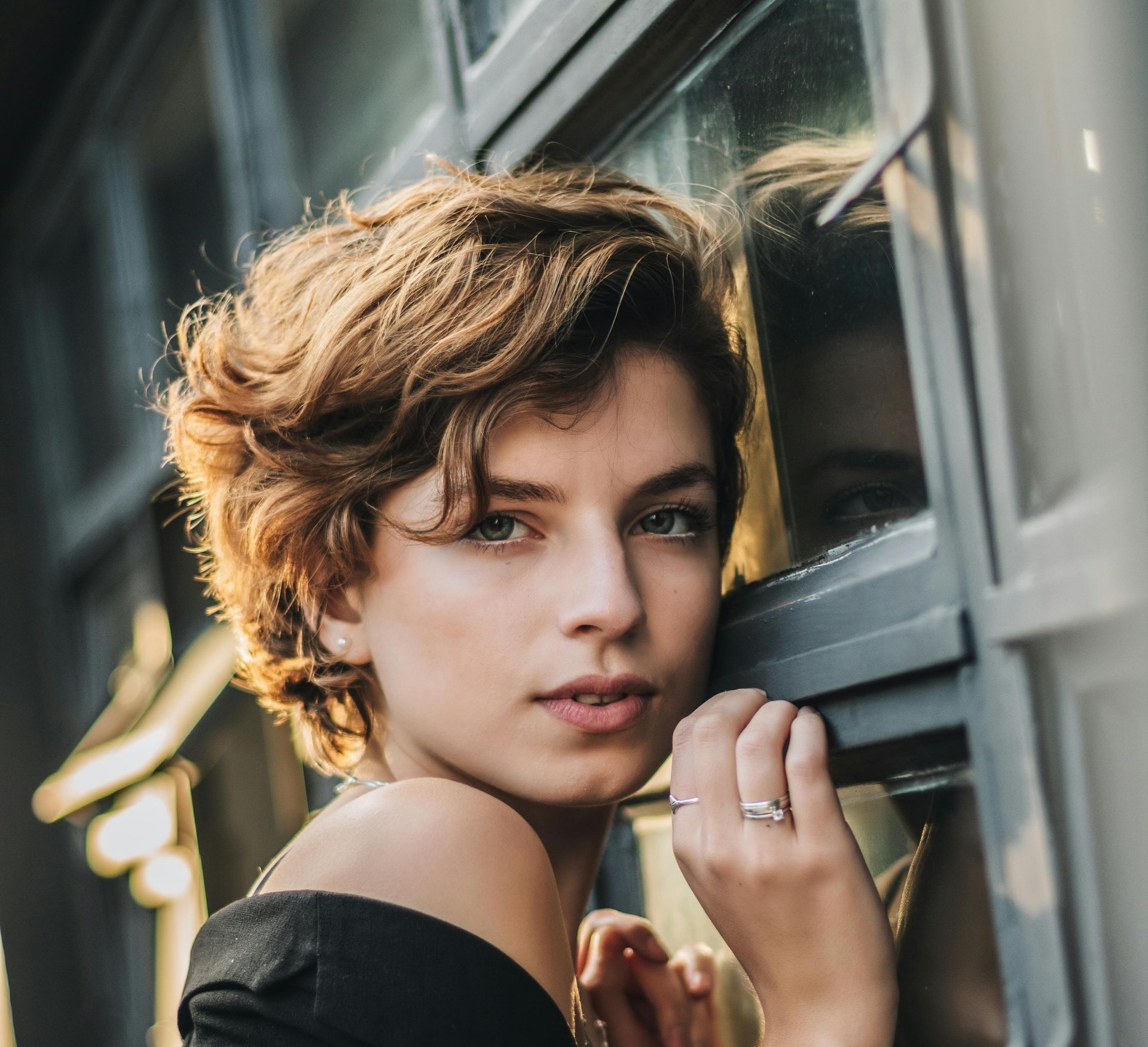 Woman with curly brown hair leans against a window, looking at the camera.