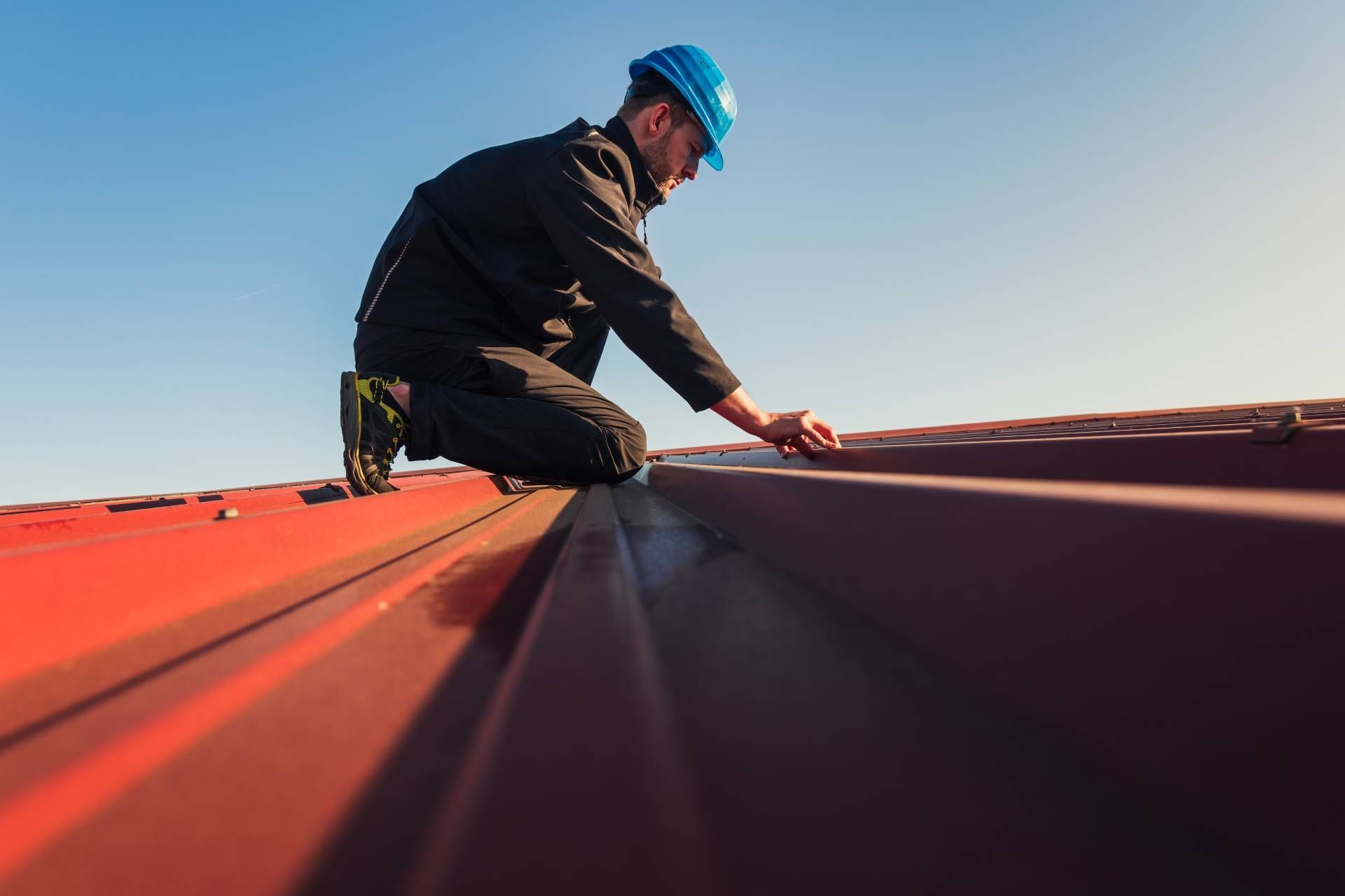 Worker in blue hard hat kneeling on a red metal roof, inspecting seams under a clear sky