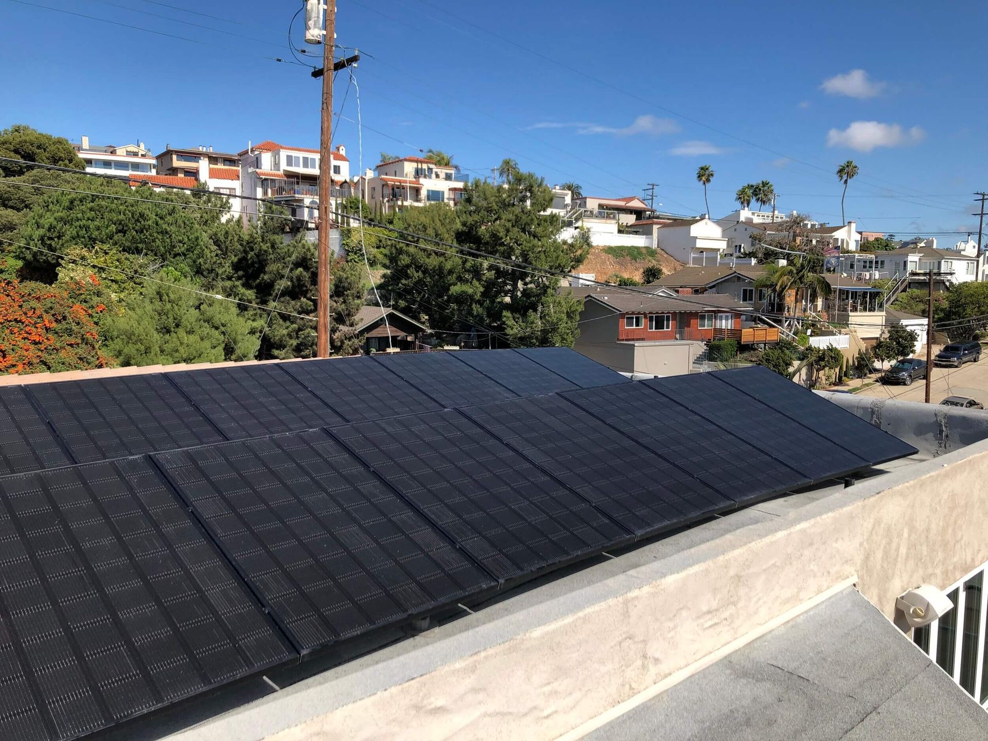 Black solar panels on a rooftop overlooking a sunny residential hillside neighborhood.