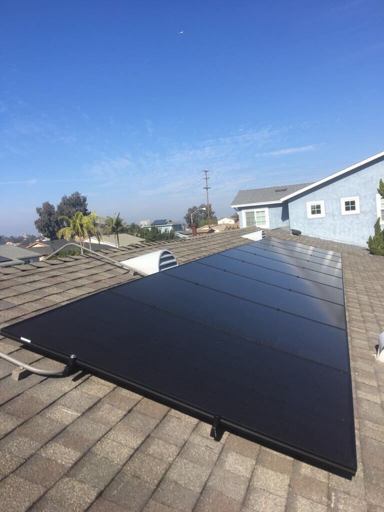 Solar panels mounted on a shingled roof beside a blue house under a clear sky.