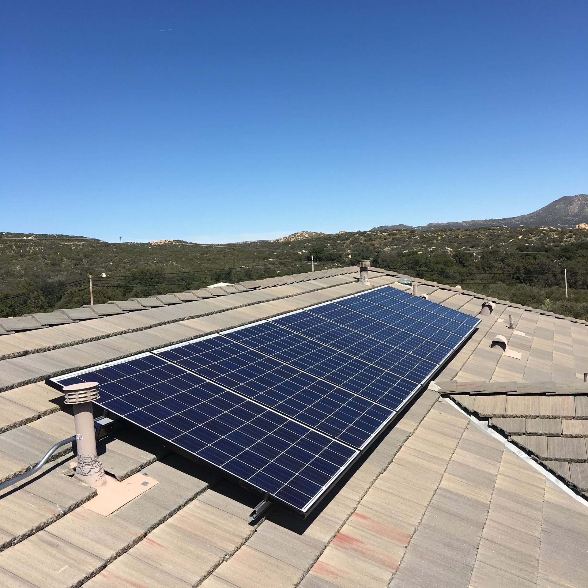 Solar panels mounted on a tiled rooftop under a clear blue sky, with hills in the background