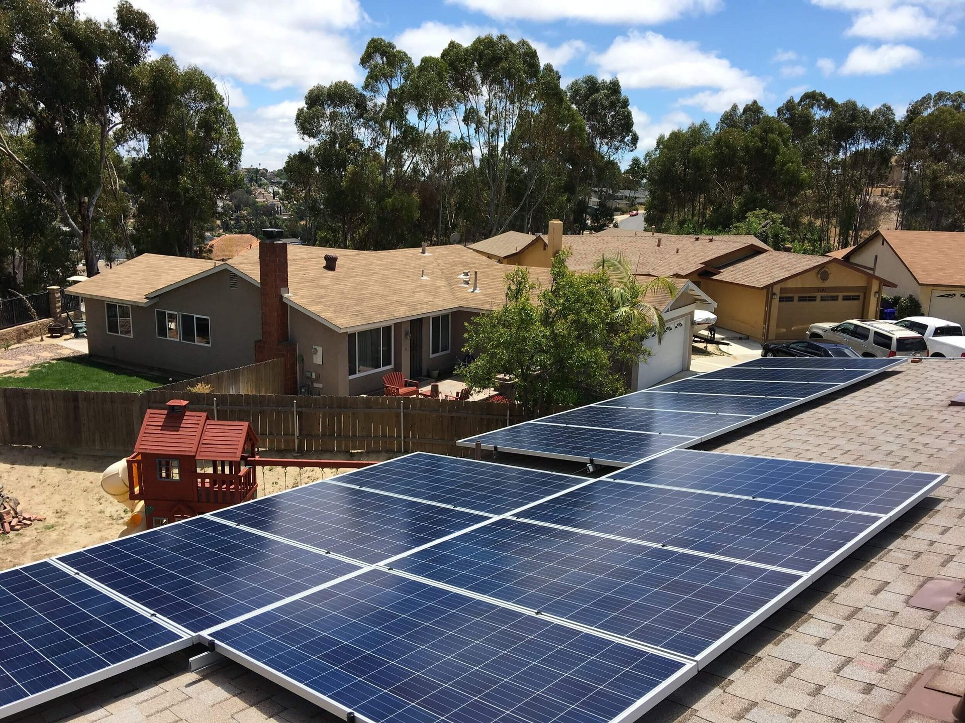 Solar panels on a rooftop overlooking suburban houses and trees under a blue sky.