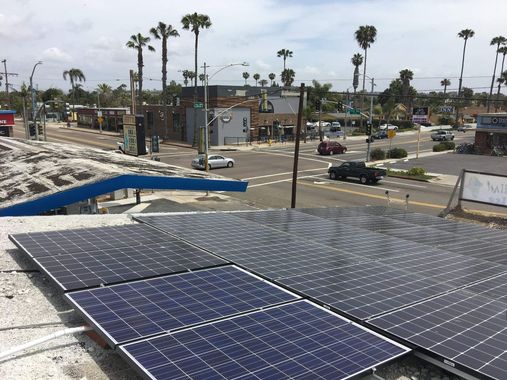Solar panels on a rooftop overlooking a palm-lined street and houses under a clear sky.