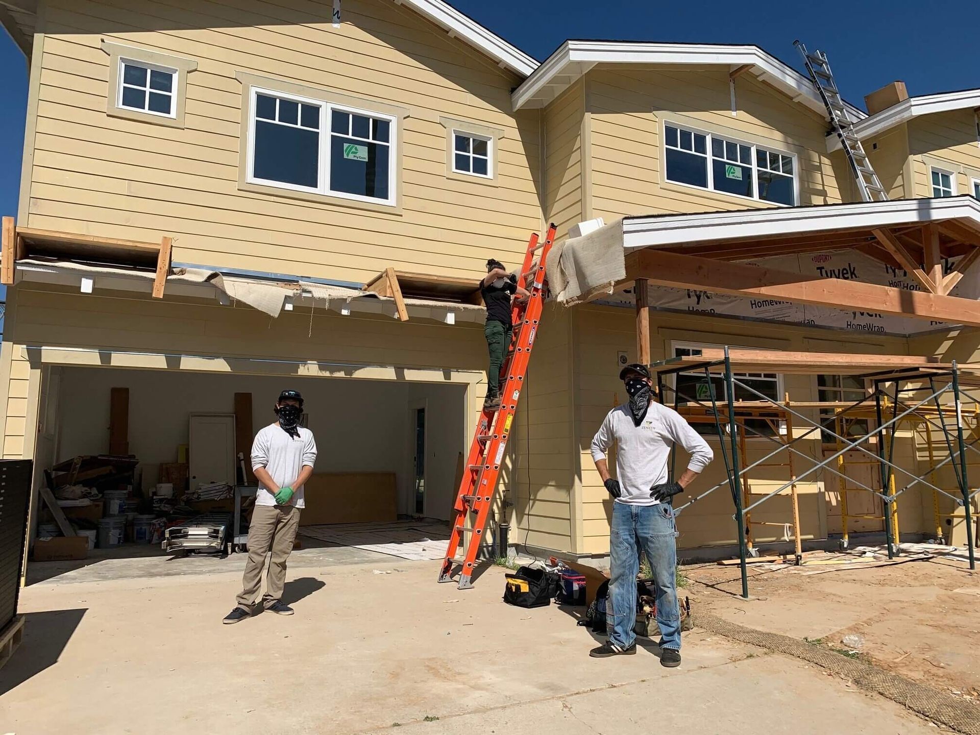 Two workers outside a beige house under construction, with scaffolding, ladders, and unfinished trim.