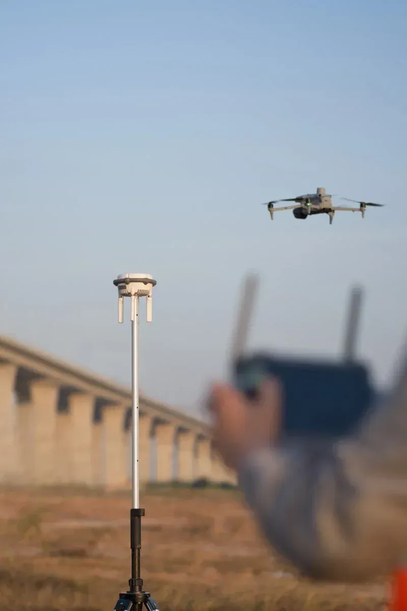 Drone hovering above a field near a person holding a controller, with a tall sensor pole in the foreground.
