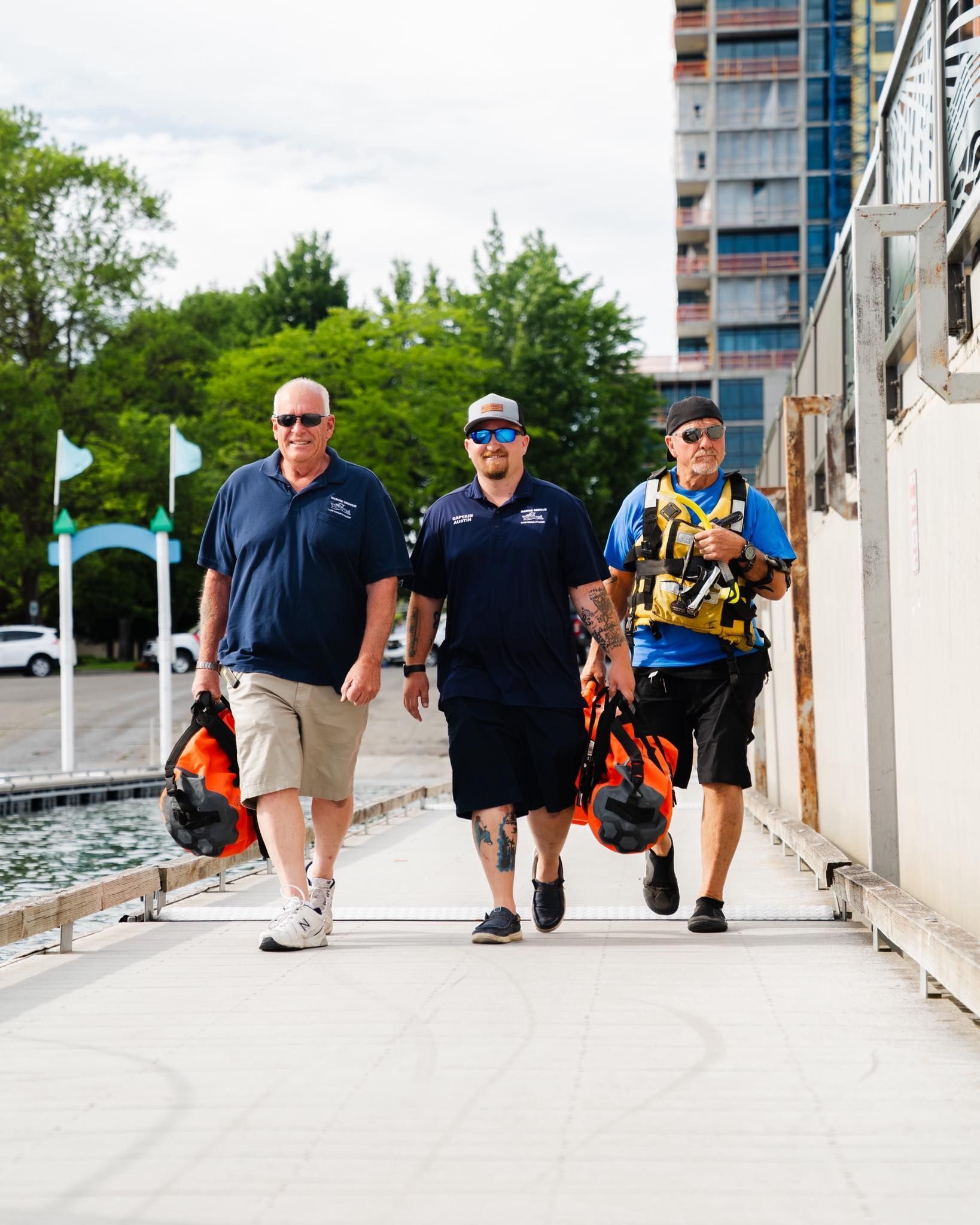 Austin Munda with two men in blue shirts and shorts walk on a dock, carrying orange bags, near water and a tall building.