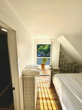 Bedroom with white walls, a bed, and a small balcony overlooking trees. Sunlight streams in.