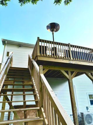 Wooden deck with stairs, attached to a white house. A patio heater sits on the deck, under a blue sky.