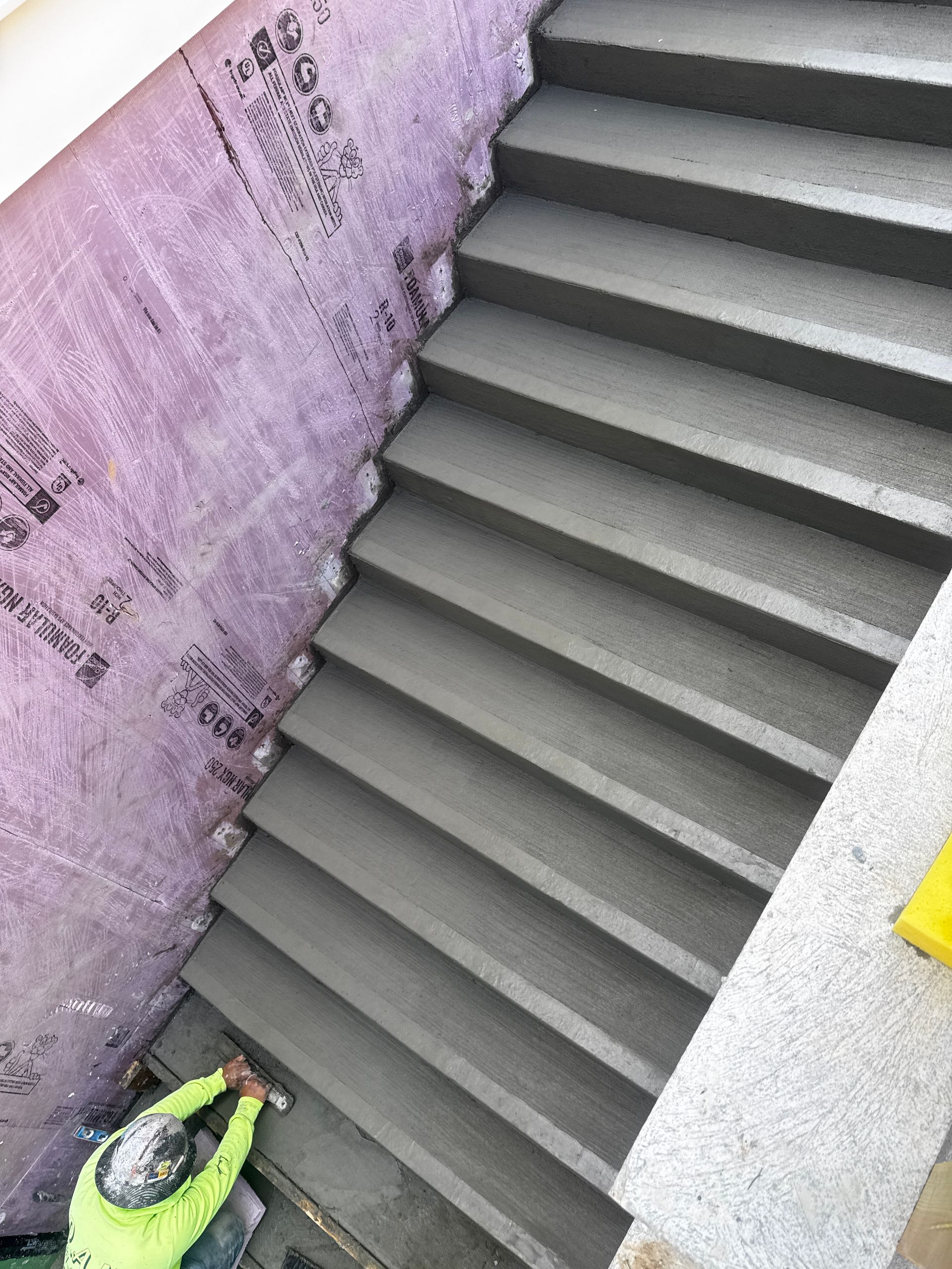 Concrete steps being constructed, next to pink insulated wall; worker in yellow vest at bottom.
