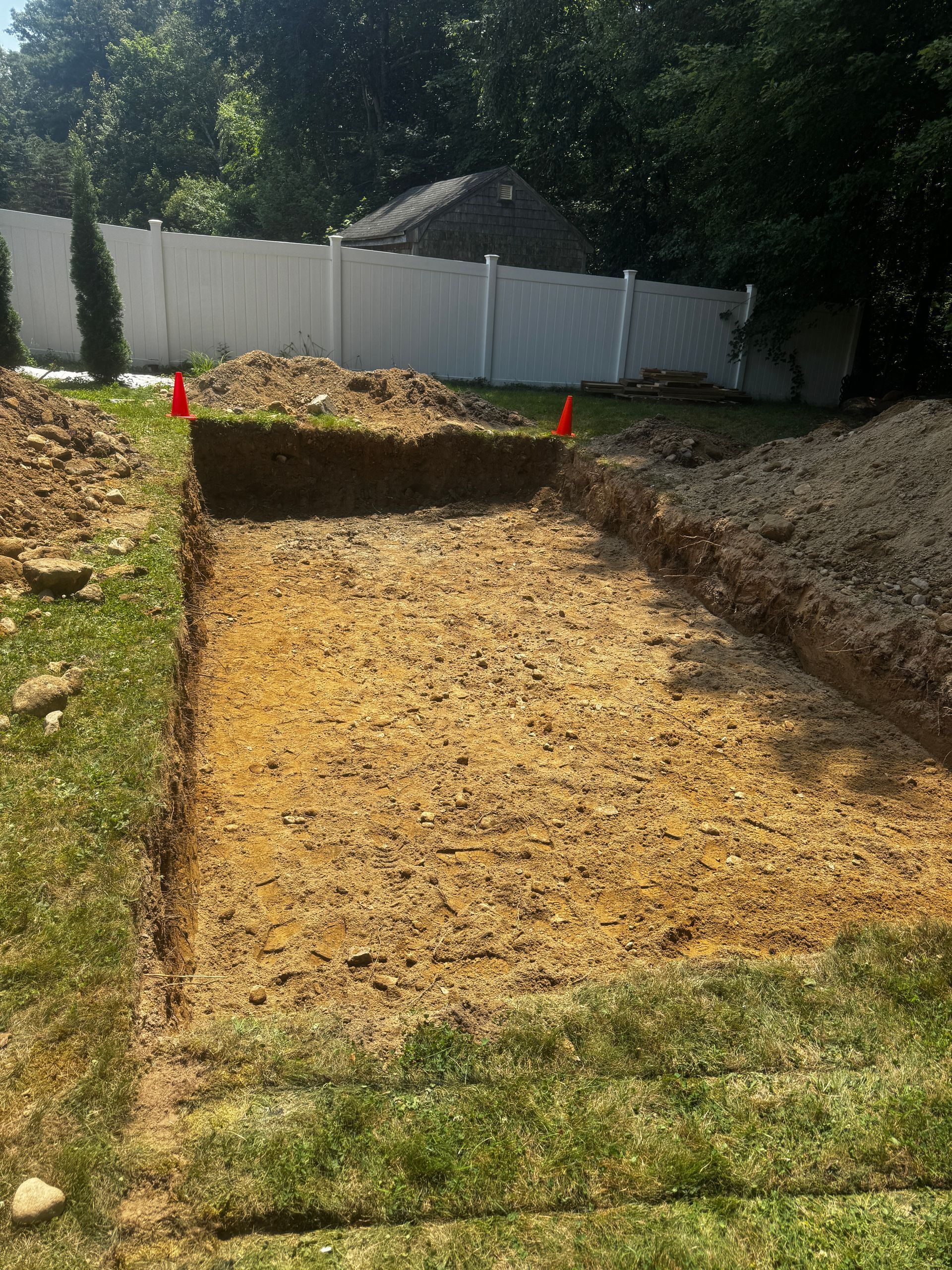 A rectangular plot dug in a grassy yard, with piles of dirt nearby. A white fence and trees are in the background.