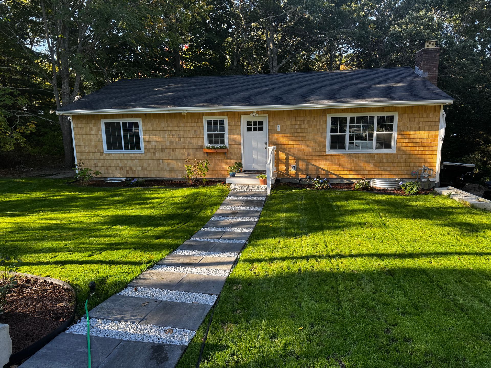 Cottage with a stone walkway leading to a white door. The house has a shingled exterior and a green lawn.