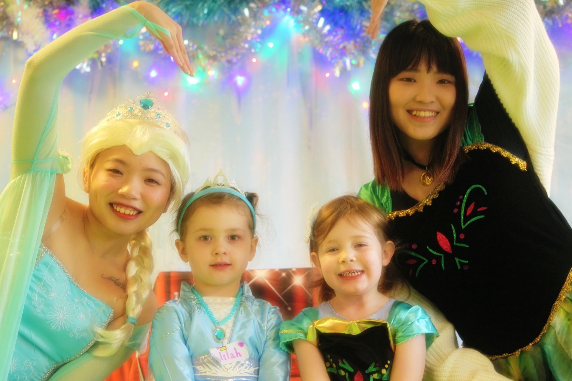 Two adults dressed as Elsa and Anna with two children dressed as Elsa and Anna, smiling. Festive background.