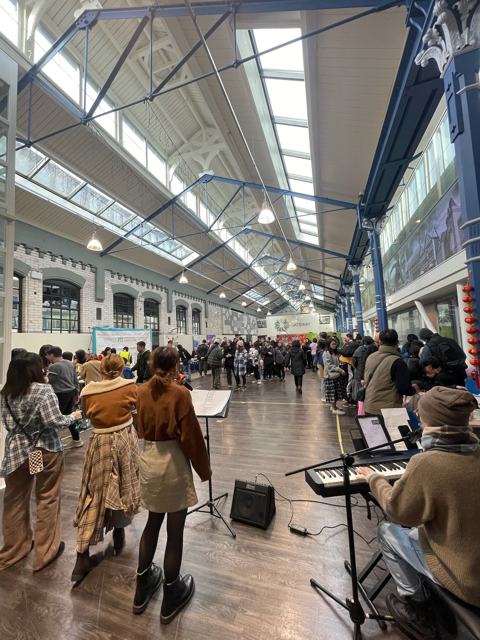 People gathered inside a large hall, some listening to a keyboard player. Sunlight streams in from above.