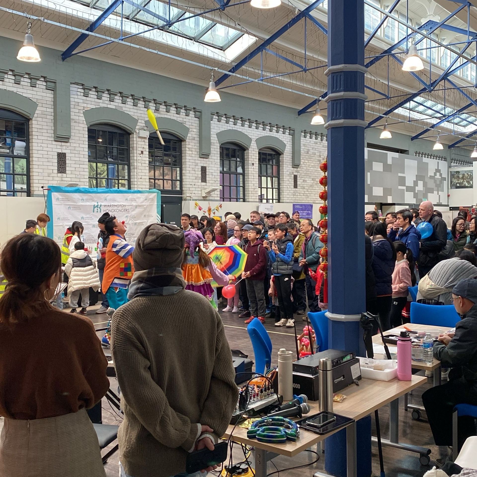 Indoor event: performers in colorful costumes, crowd watching, tables with equipment, in a large hall with windows.