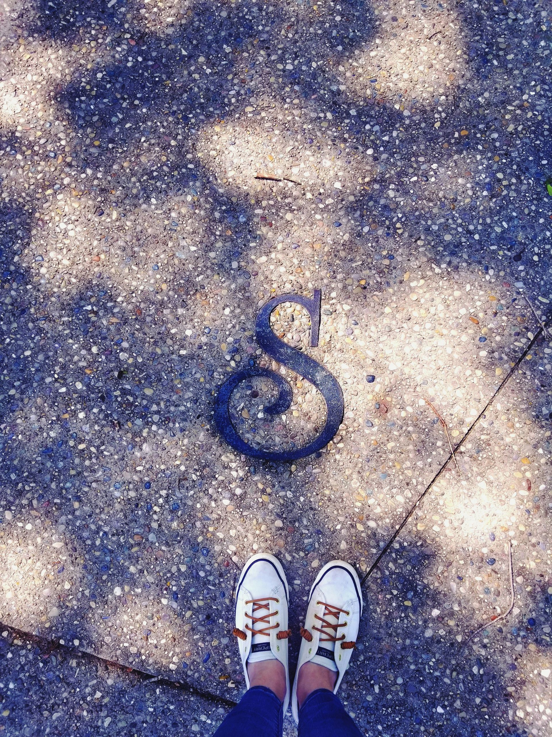 Person's feet in white sneakers standing over a dark ampersand symbol painted on concrete.