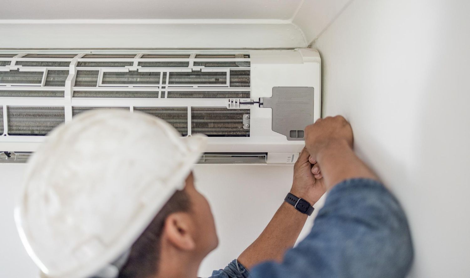 Person in a hard hat repairing a wall-mounted air conditioning unit.