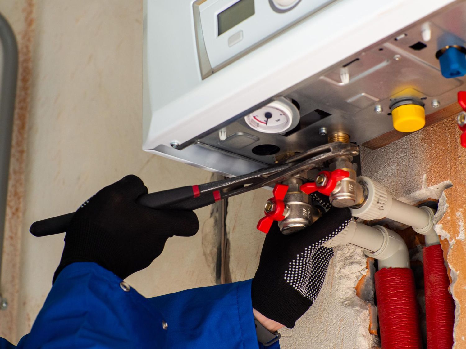 A person in gloves using a wrench on pipes connected to a boiler, in a wall.