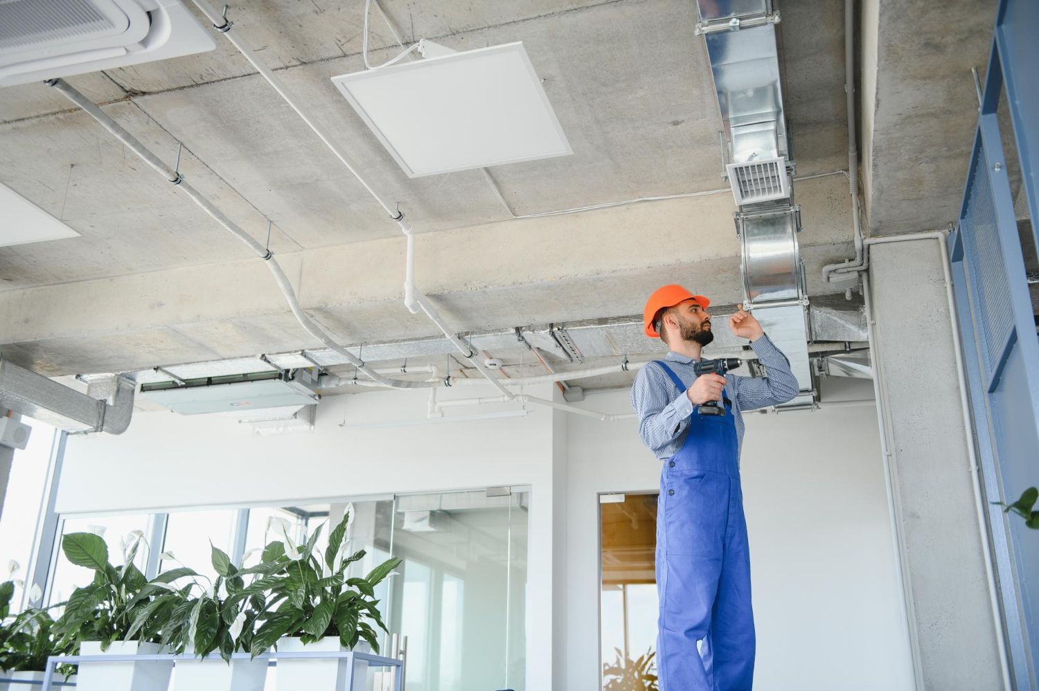 HVAC technician in blue overalls and orange hard hat working on ductwork in a building.