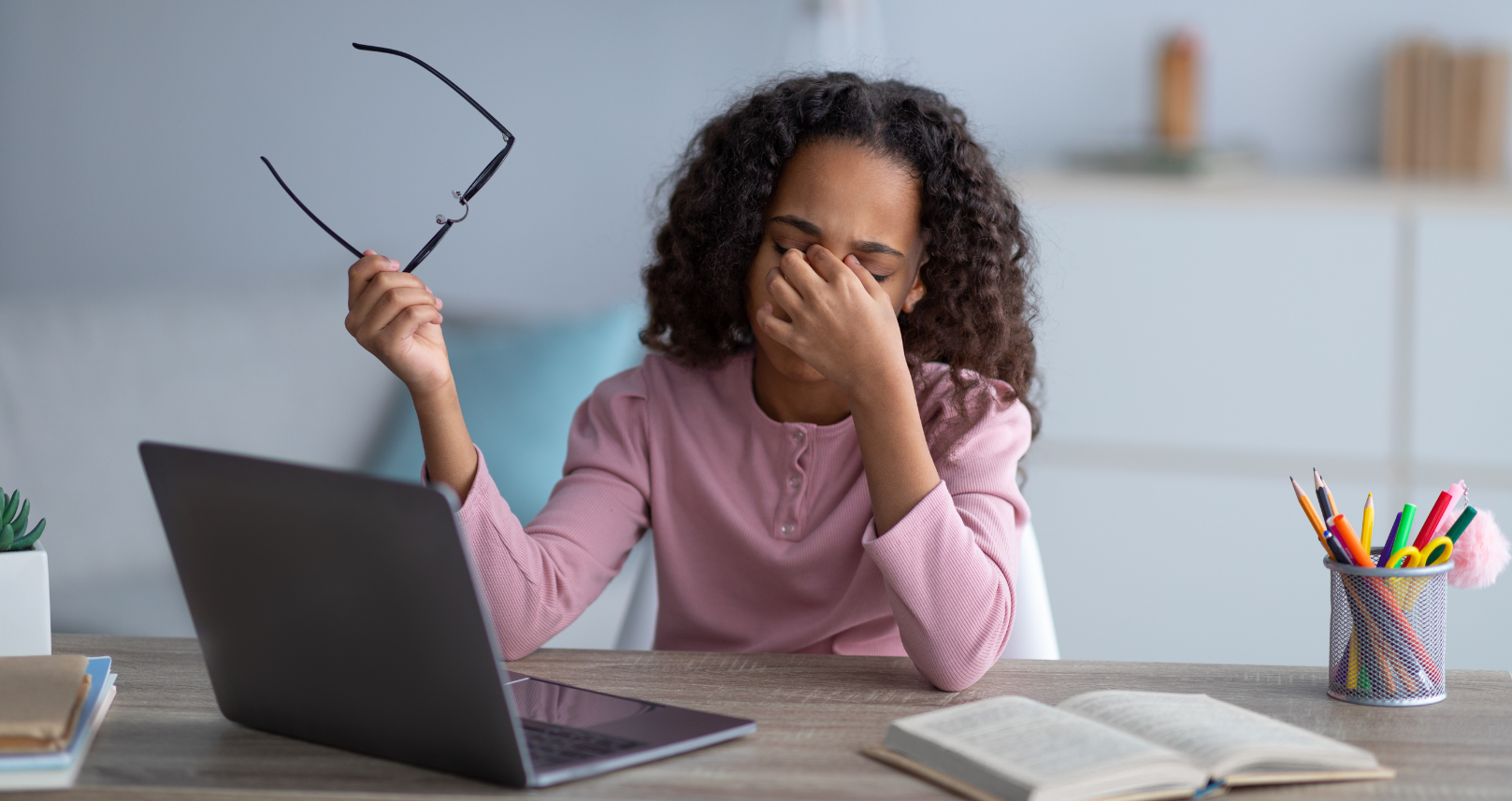Person at a desk rubbing their eyes while holding glasses, with a laptop, notebook, and pencil cup nearby.